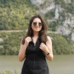 A fashionable woman in sunglasses smiling by a picturesque lake with mountains in the background.